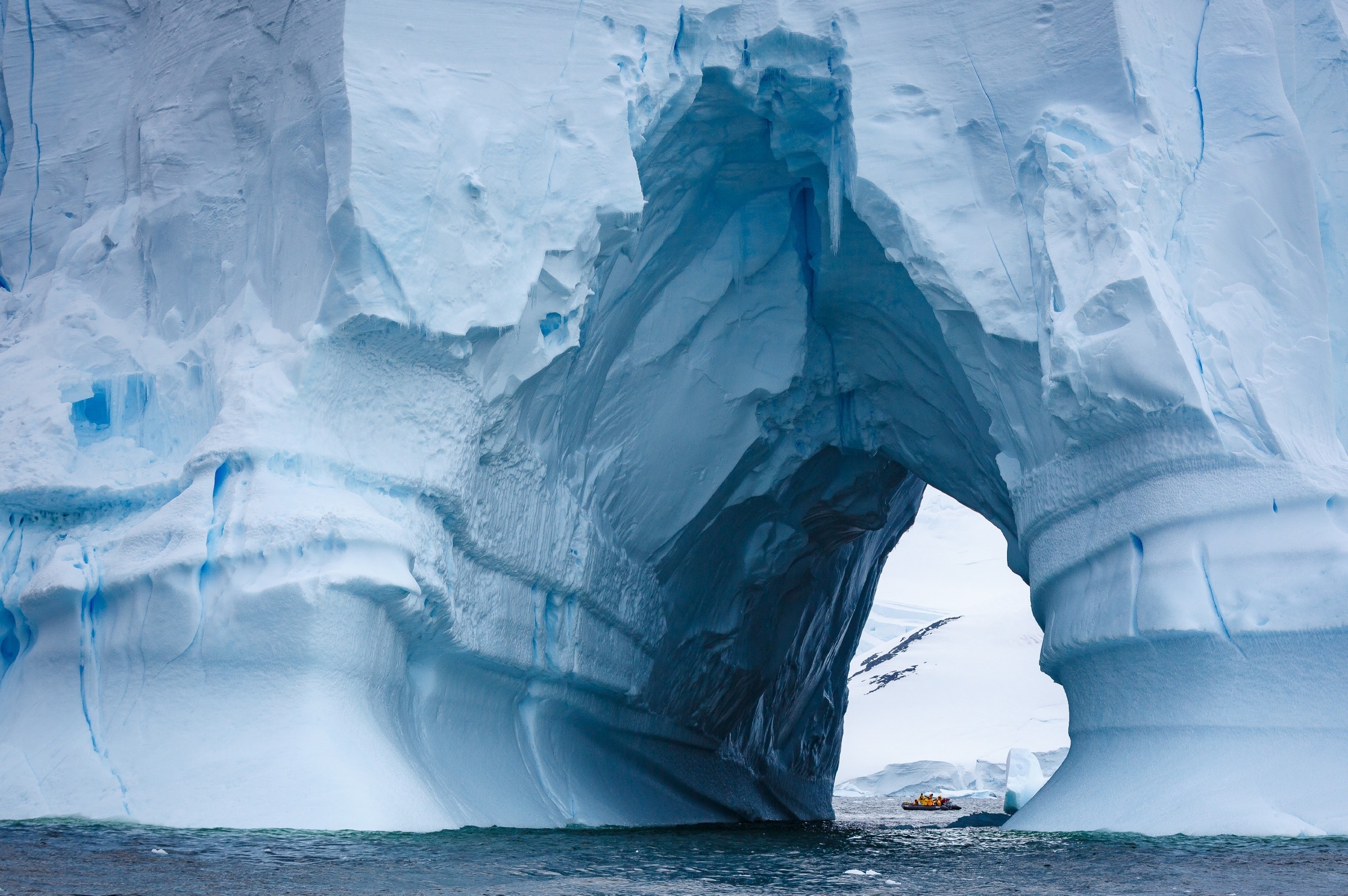 A mammoth iceberg fills the entire frame of the shot, with a natural tunnel through it revealing a zodiac boat with tourists on the other side.