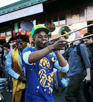 A young man with a bright green baseball hat and blue t-shirt plays a trombone in the street with other revellers, some donning Venetian masks, others feathers in their hair