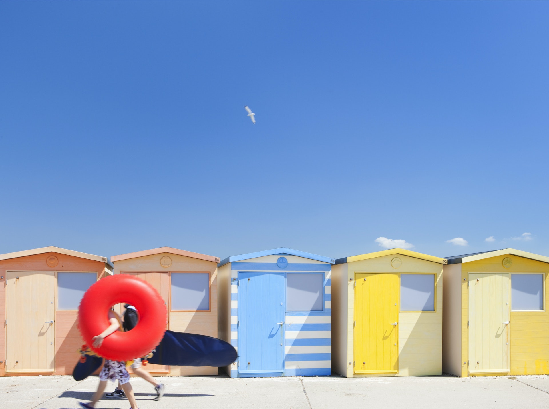 People carrying inflatables walk past colorful beach huts on the English coast