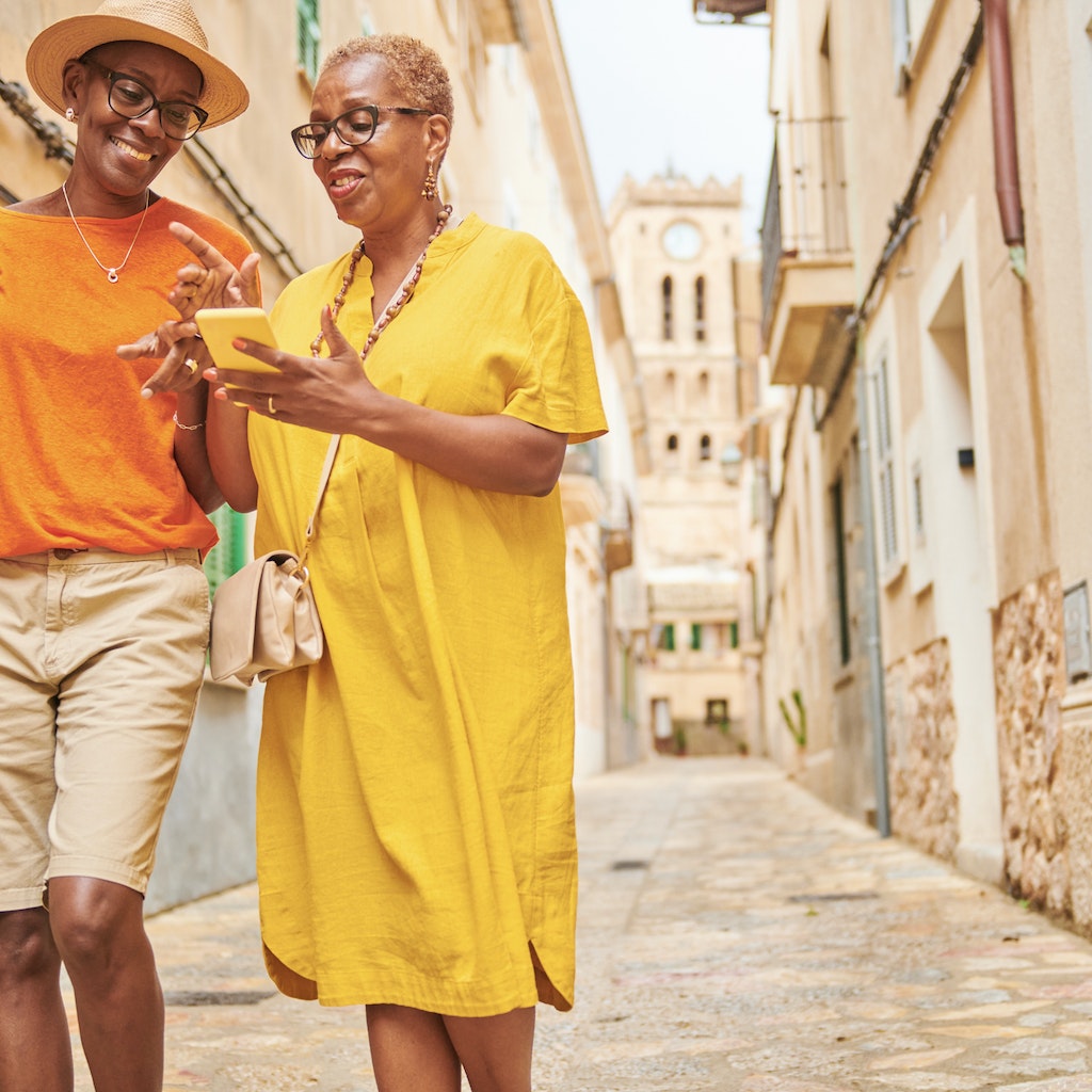 Using a mobile app on their smart phone to guide their way around a traditional old town in Majorca Spain.
1438234950
Two older black women walking along a cobbled street in Spain while looking at their smartphone and laughing