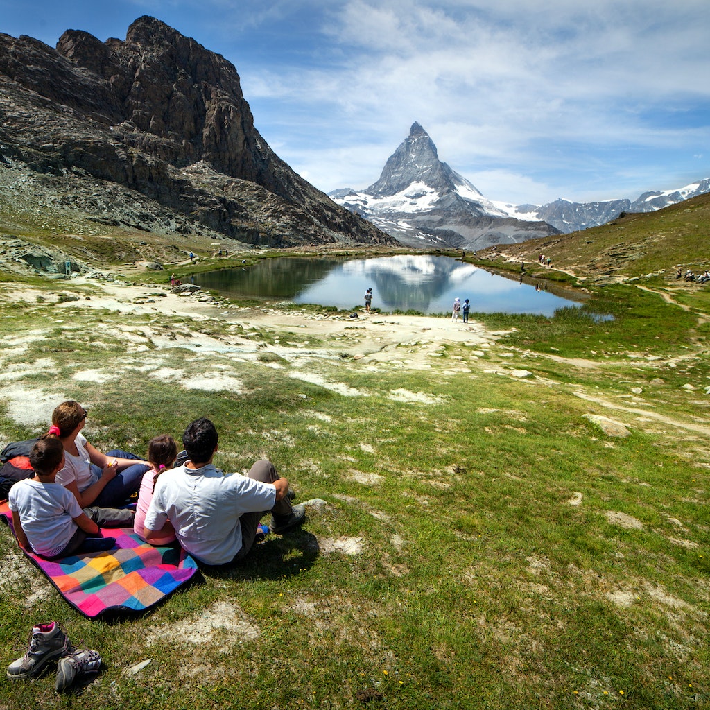 Summertime in Switzerland. A family of four is having fun relaxing on the lawn in front of the Matterhorn. The mountain peak is reflecting into the water of a mountain lake. Horizontal composition, Outdoor experience.
915075652