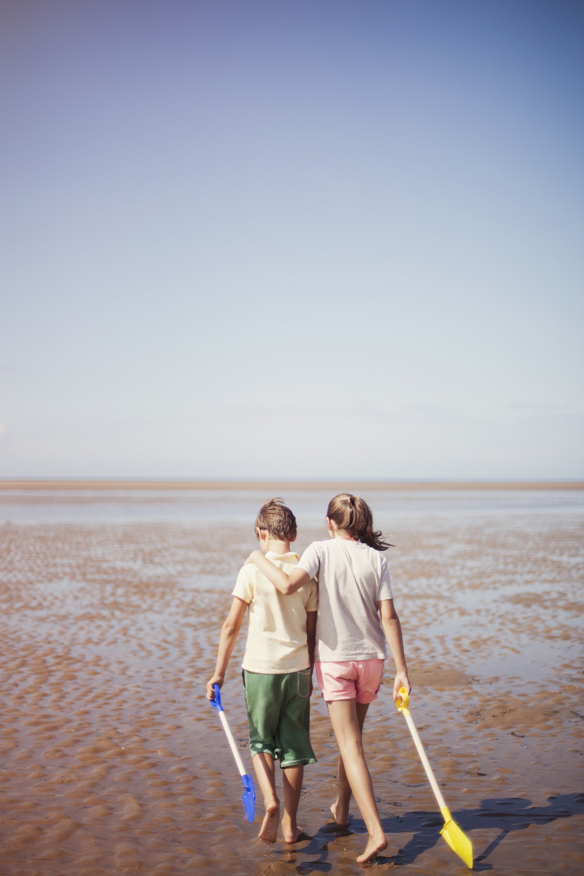 Brother and sister with shovels hugging and walking in wet sand on sunny summer beach below blue sky
