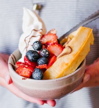 A bowl heaped with strawberries, blueberries, vegan cream and a waffle; the bowl cupped by a pair of hands