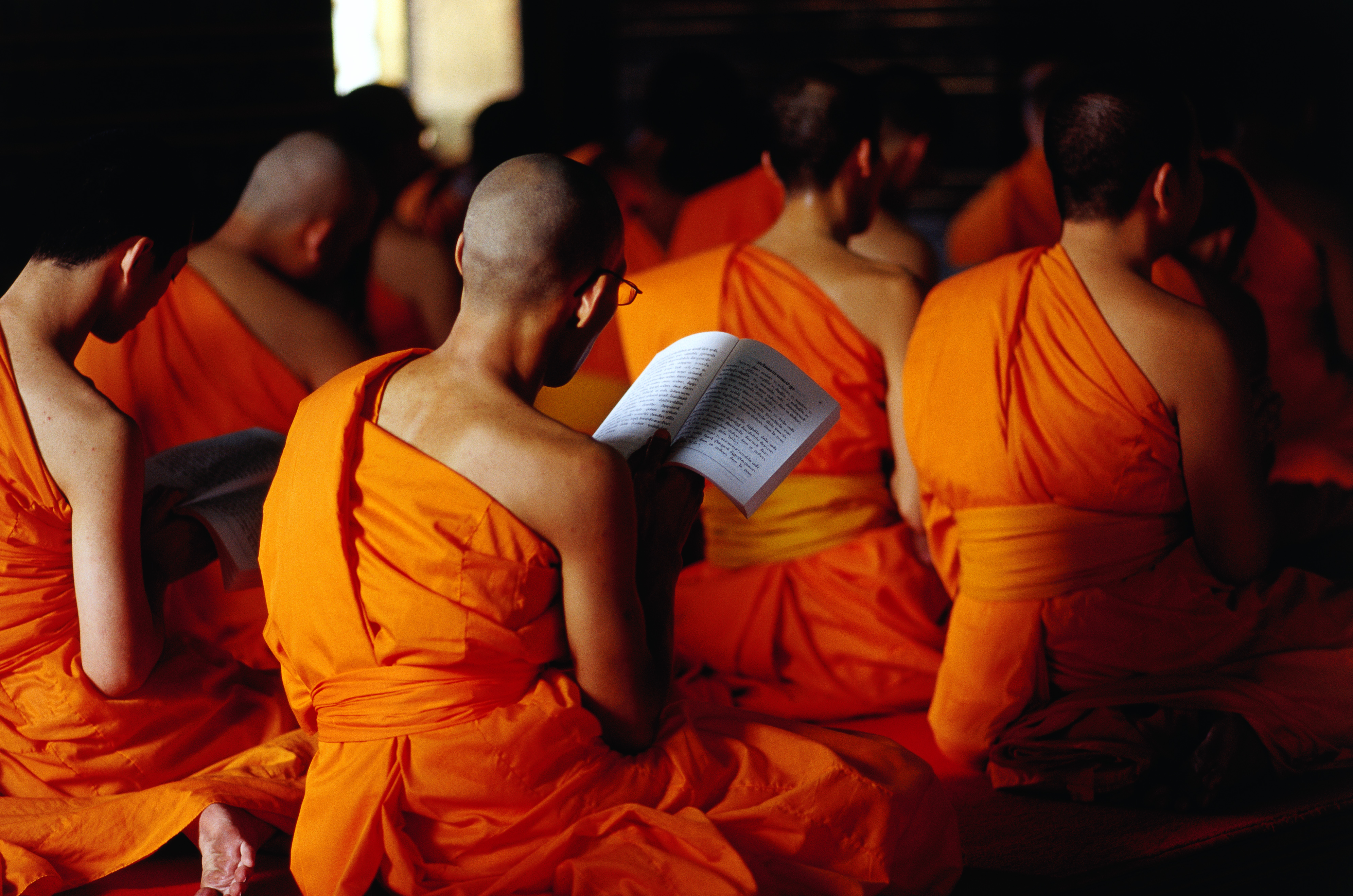 Shaved-headed monks sit cross legged at morning prayers in sermon hall at Wat Pho, some holding prayer books.