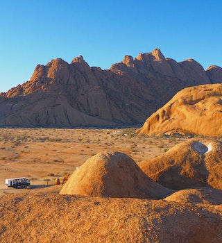 A large overland truck looks much like a piece of lego as it sits on the red Namibian soil surrounded by massive, bulbous rock monoliths at Spitzkoppe @ Sarah Reid / Lonely Planet