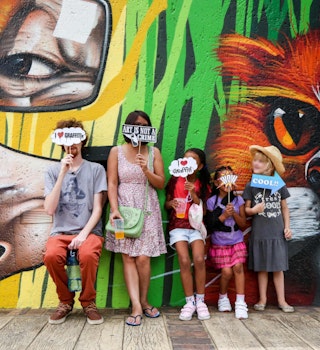 Two adults and three children stand in front of a wall covered in street art. The man holds a small cloud-shaped sign that covers his face and reads 'I heart graffiti'. The woman holds a similar sign that reads 'Art is not a crime'. The children hold similar sings, including ones that say 'Wow' and 'Cool'.© Heather Mason / Lonely Planet
