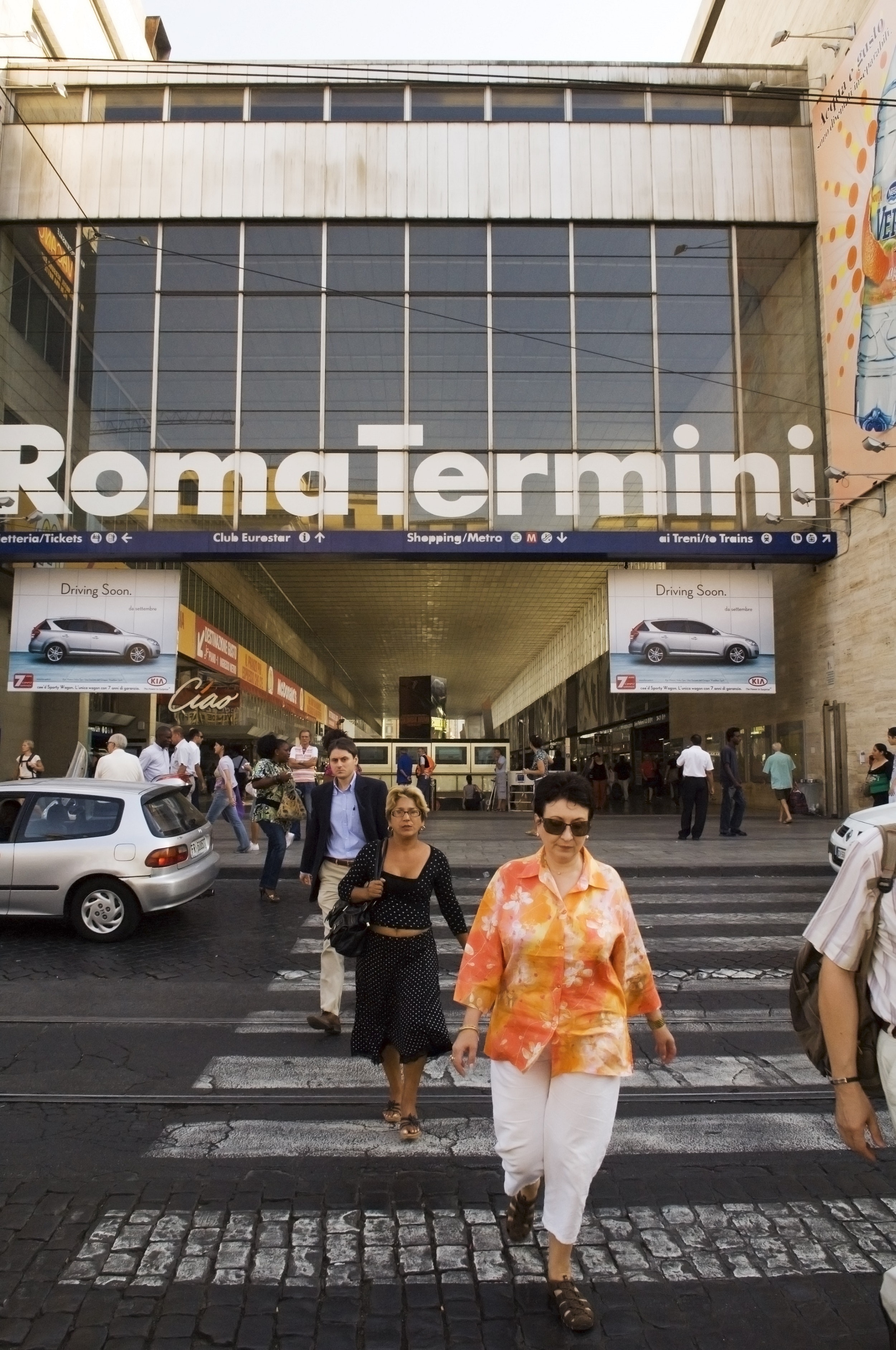 People walk across a zebra crossing in Rome towards the camera; behind them is a large building with huge glass windows with large white letters that read: Roma Termini