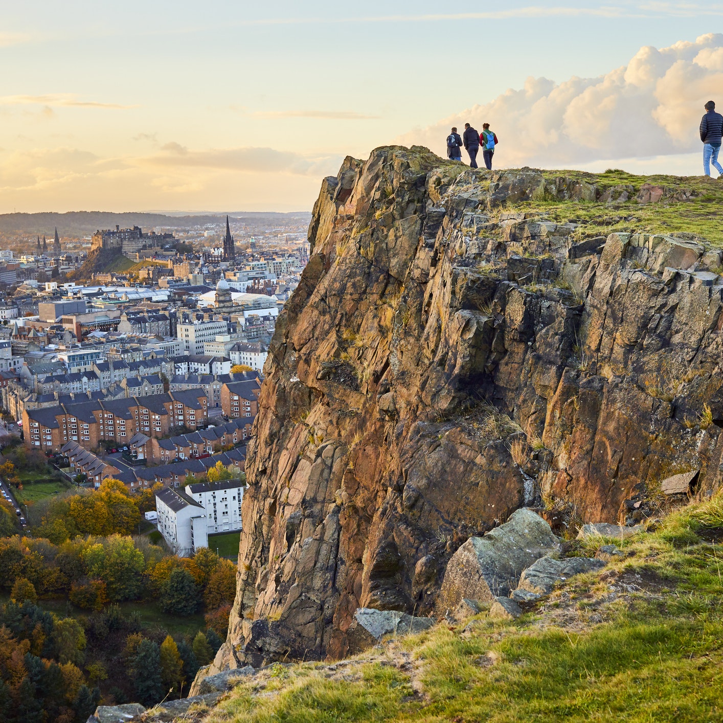 Group of people walking along cliff edge looking at city views. Edinburgh Castle in the distance