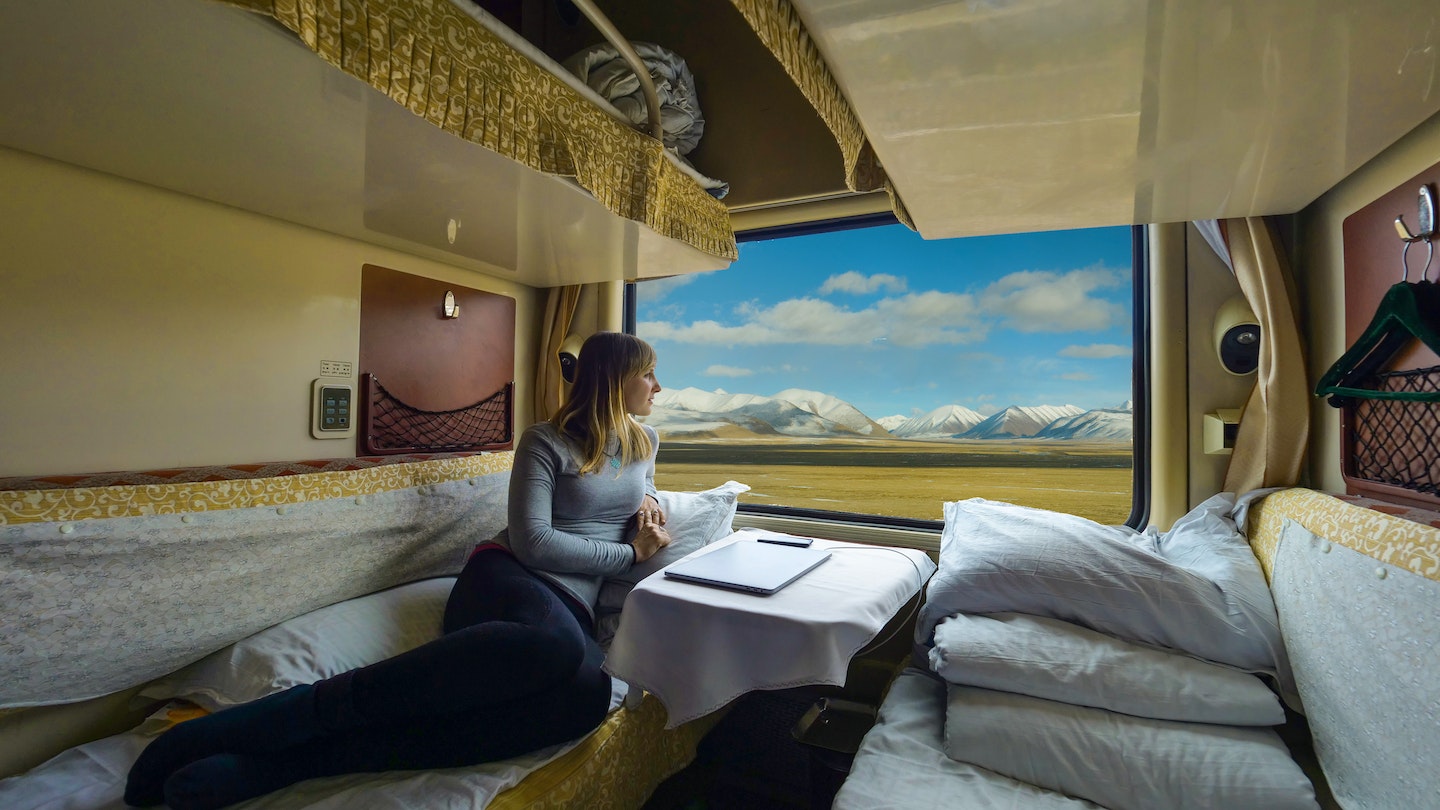 Young woman lies on the bed and looks through the window of the sleeper train crossing picturesque Tibet. Female tourist observing the landscape while traveling along the Trans-Himalayan railway.