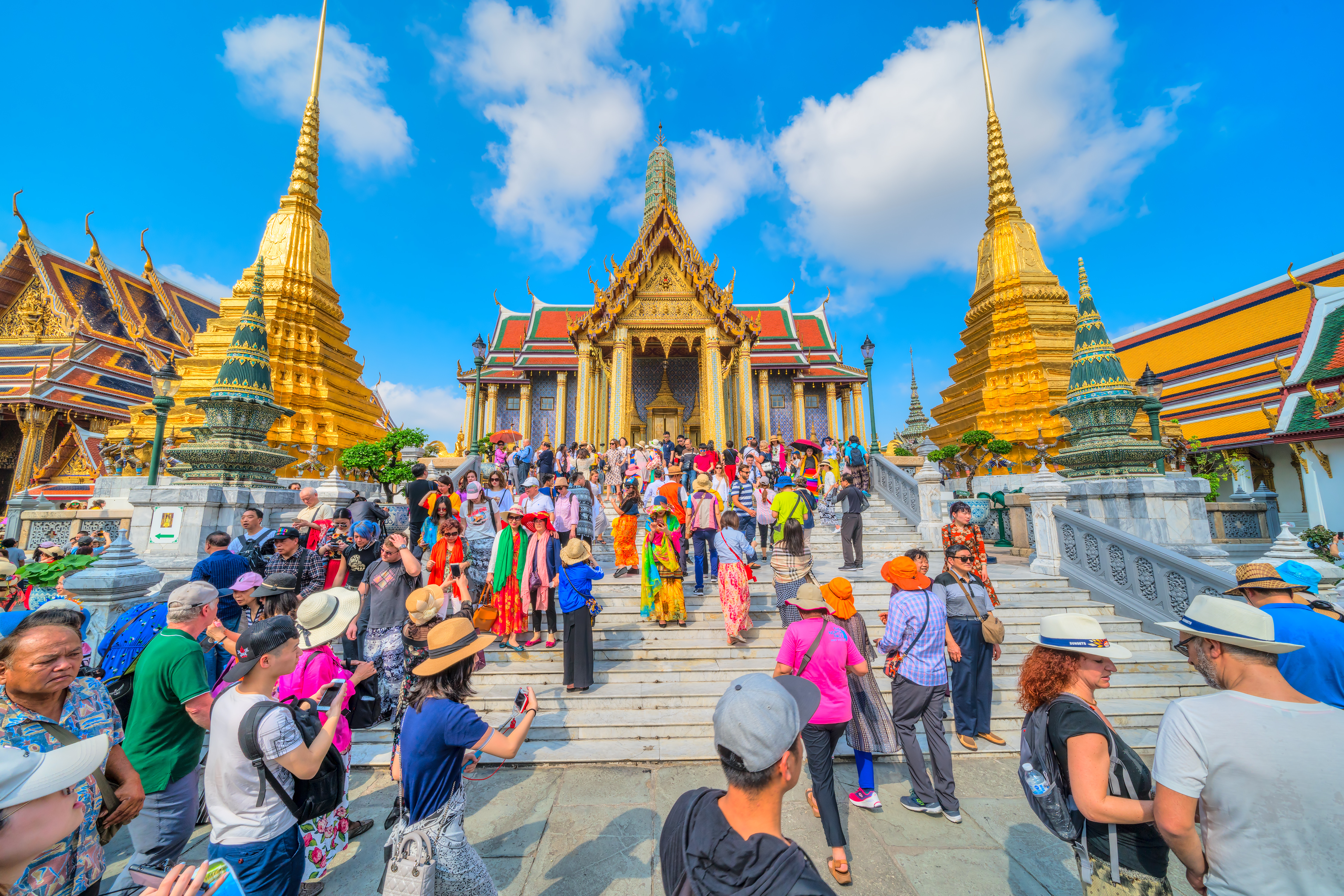 People mill around the complex of the Grand Palace in Bangkok as its golden spires pierce the blue skies.