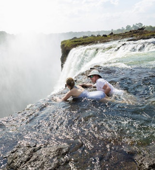 Man and a girl, father and daughter in the water of the Devil's Pool on the edge of Victoria Falls, mist rising from the falling water.